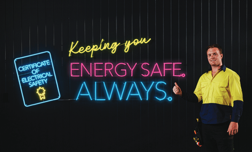 A graphic depicting neon lights to represent a certificate of electrical safety and put forth the message "Keeping you Energy Safe. Always.", with a tradesperson standing to the right of the neon text as an expression of approval.