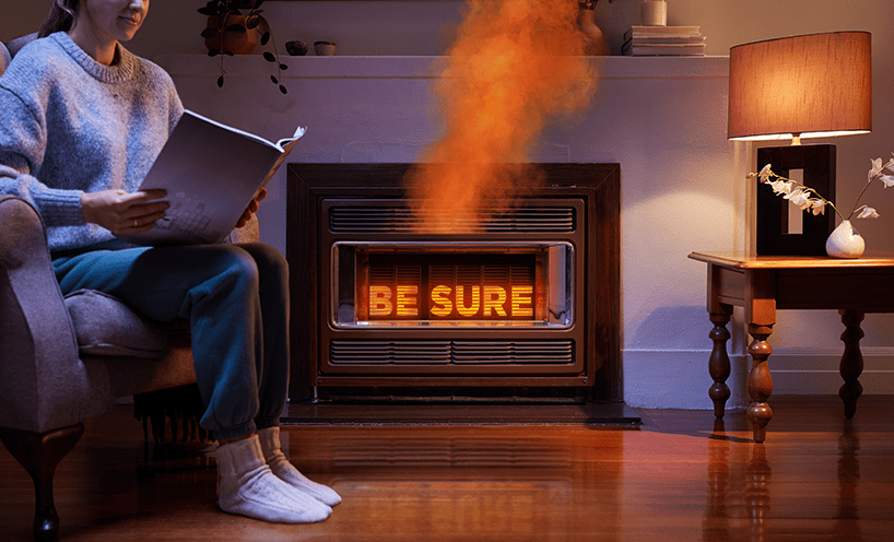 A person sits, reading, with a gas heater in the background that's emitting orange fumes – representing the invisible danger of carbon monoxide – and the heater's grill reads 'Be Sure' via an orange glow.