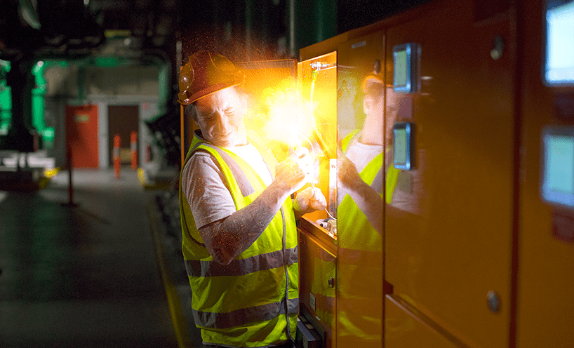 An electrician winces as electricity – represented as a burst of light with sparks – erupts from a panel they are working on.